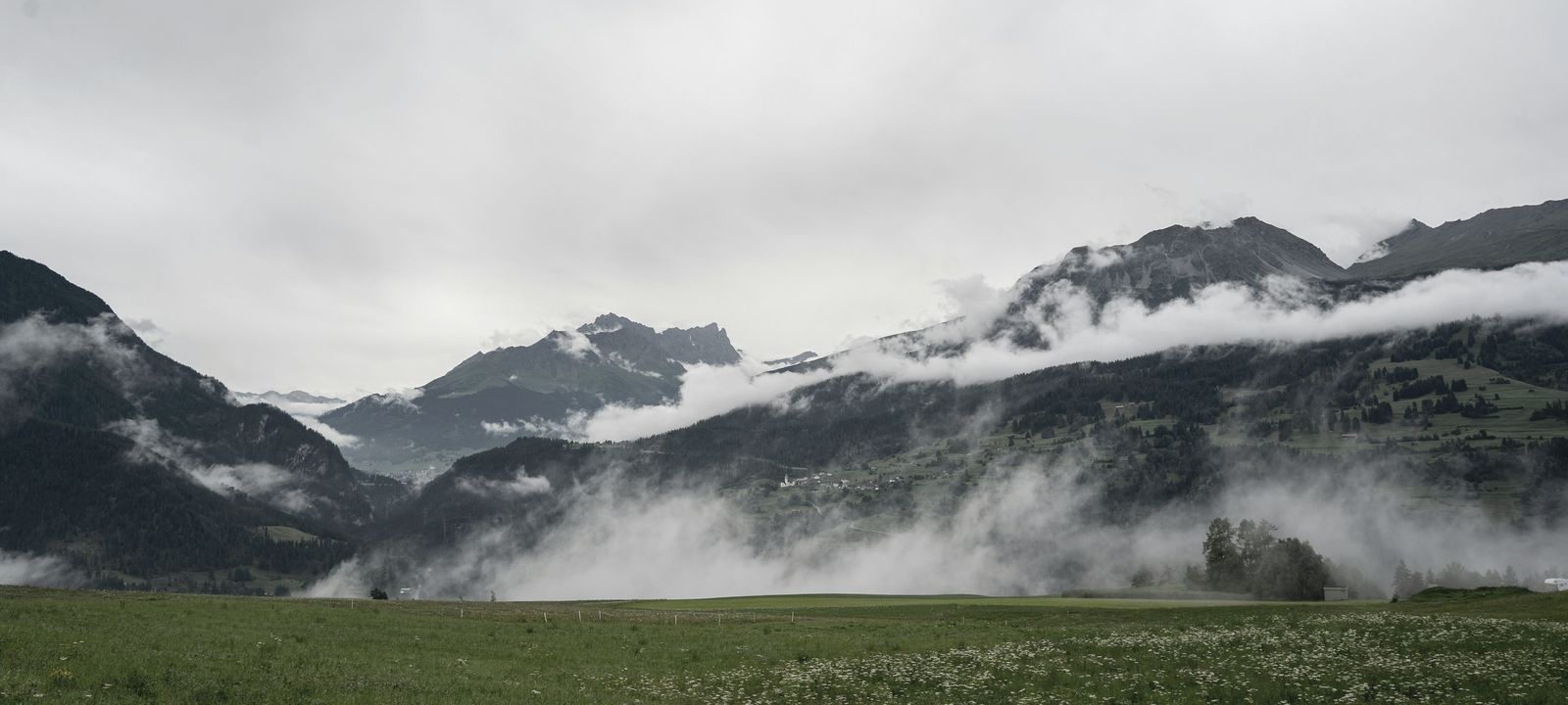 A farmhouse in a green meadow under a band of low cloud, mountains behind