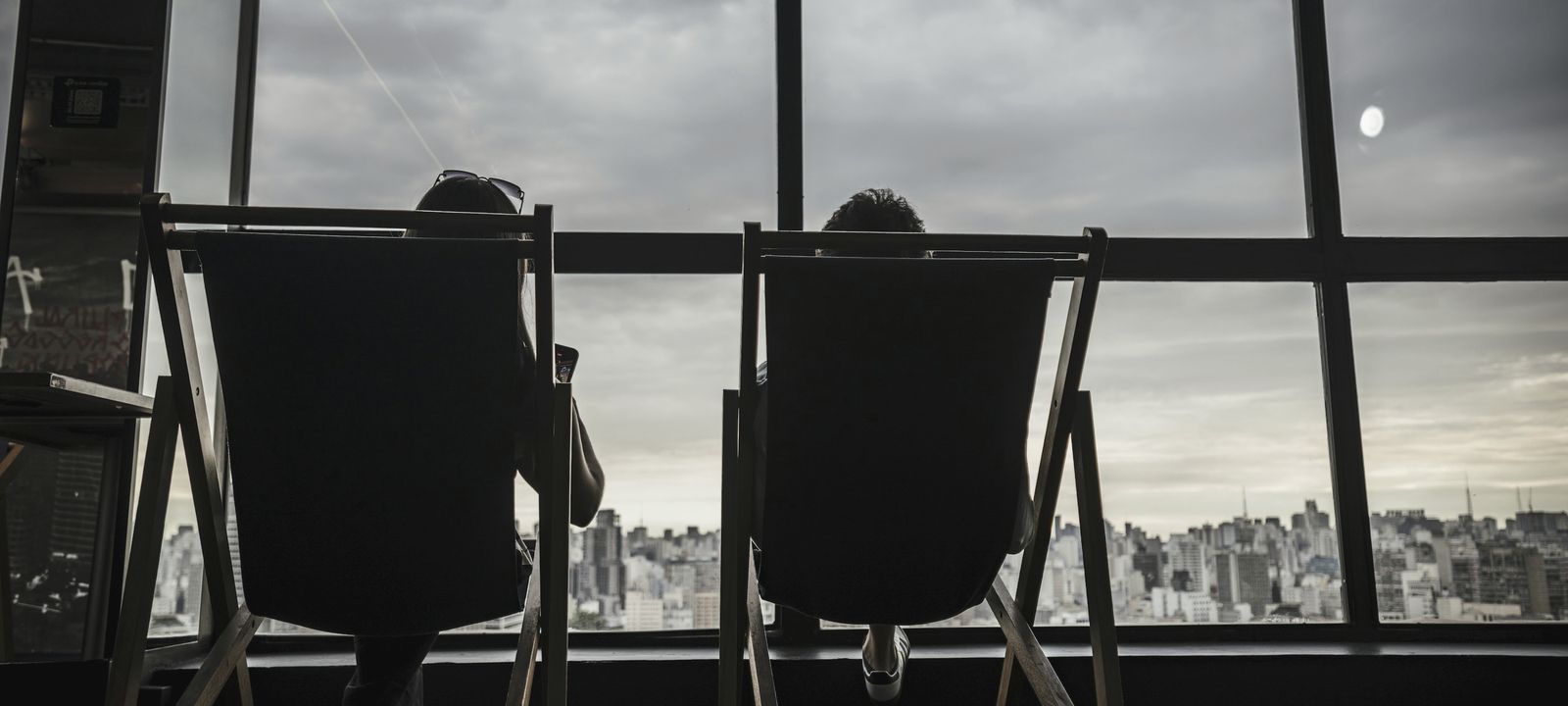 Two people in deck chairs, seen from behind, looking out through a tall window at a city under cloud