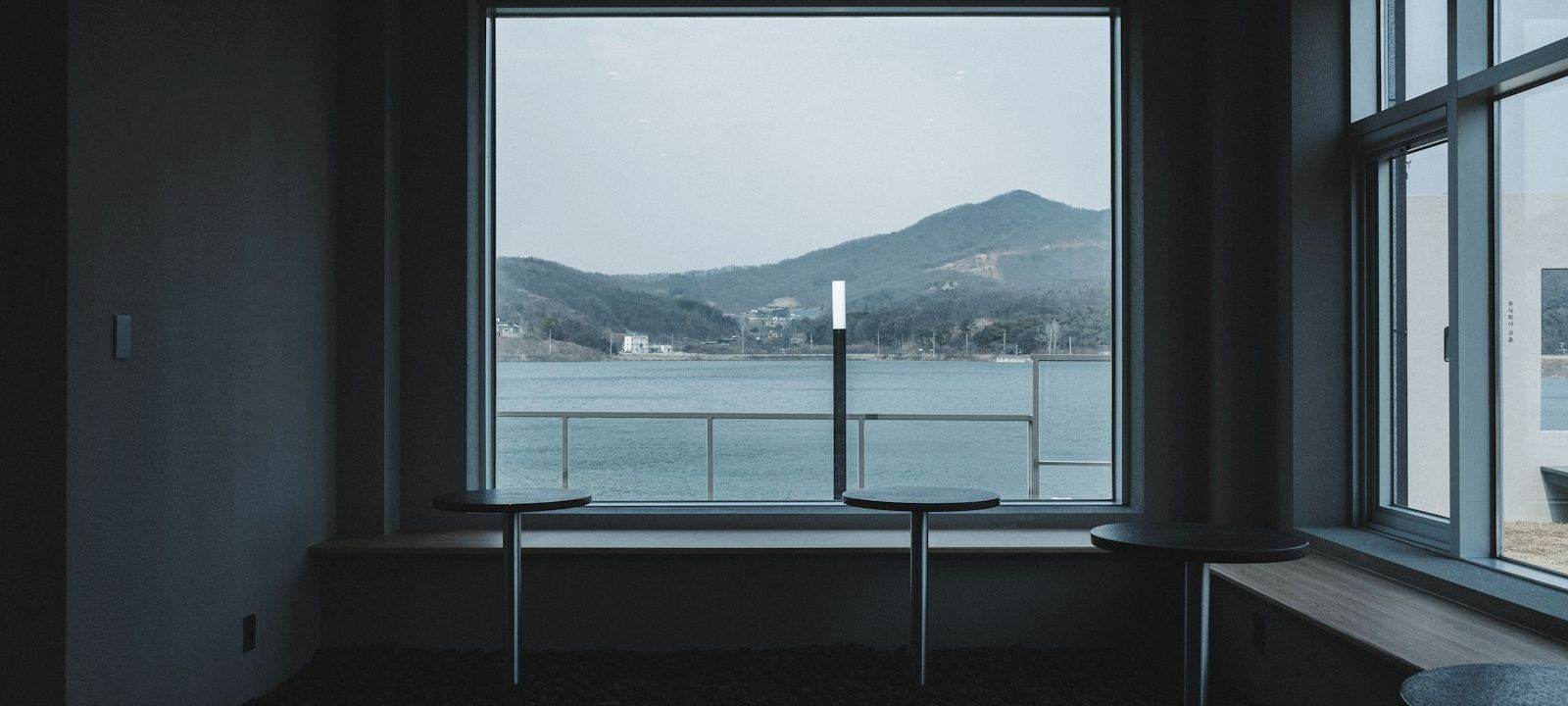 A quiet room with small tables along a window seat, looking out over water to wooded hills